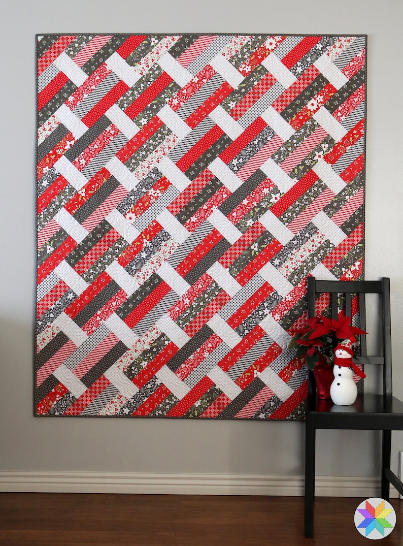 May include: A red, white, and gray patchwork quilt with a geometric pattern. The quilt is hanging on a gray wall and is framed by a white baseboard.