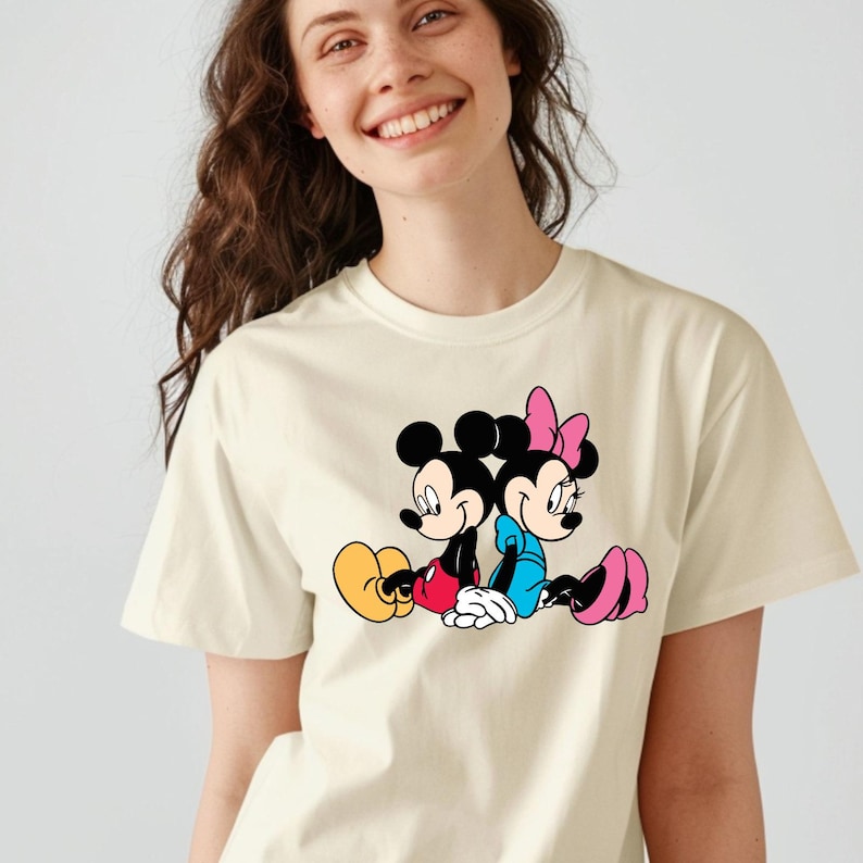 May include: A cream-coloured t-shirt with a graphic of Mickey Mouse and Minnie Mouse sitting back-to-back. Mickey wears red shorts and yellow shoes, Minnie a blue dress and pink shoes. The cartoon characters are in a classic pose.