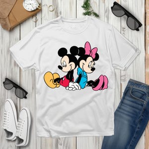 May include: White t-shirt with a cartoon illustration of Mickey and Minnie Mouse. Mickey is in red shorts and yellow shoes, Minnie in a blue dress and pink shoes. The image is a classic cartoon design, perfect for fans of the characters.