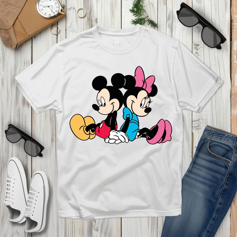 May include: White t-shirt with a cartoon illustration of Mickey and Minnie Mouse. Mickey is in red shorts and yellow shoes, Minnie in a blue dress and pink shoes. The image is a classic cartoon design, perfect for fans of the characters.