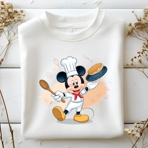 Puede incluir: Sudadera blanca de cuello redondo con un diseño de Mickey Mouse como chef. Mickey lleva un gorro de chef, un pañuelo rojo y zapatos amarillos, sosteniendo una cuchara de madera y un pastel. Fondo con ilustraciones temáticas de repostería.