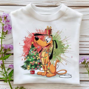 May include: White sweatshirt featuring a cartoon dog with a Christmas tree and wrapped in lights. The dog is orange and yellow with black ears. A red and green watercolor background adds a festive touch. A gift box sits on the dog's head.