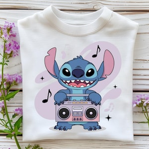 May include: White sweatshirt with a cartoon character in blue, large ears, and a wide grin, holding a pink and grey boombox. Musical notes and stars surround the character against a light purple backdrop. The sweatshirt is folded.