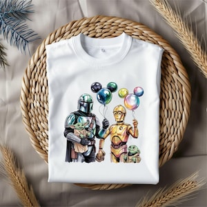 May include: White t-shirt featuring a colorful illustration of characters from the Star Wars franchise. The design includes The Mandalorian, C-3PO, and Grogu holding balloons. The shirt is displayed on a woven surface.