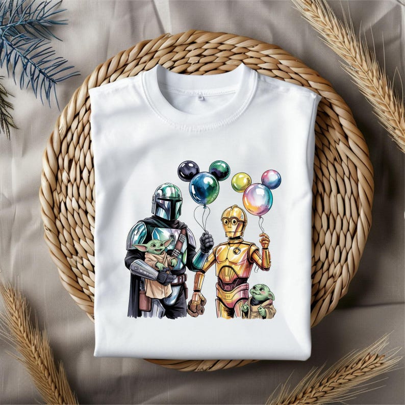 May include: White t-shirt featuring a colorful illustration of characters from the Star Wars franchise. The design includes The Mandalorian, C-3PO, and Grogu holding balloons. The shirt is displayed on a woven surface.