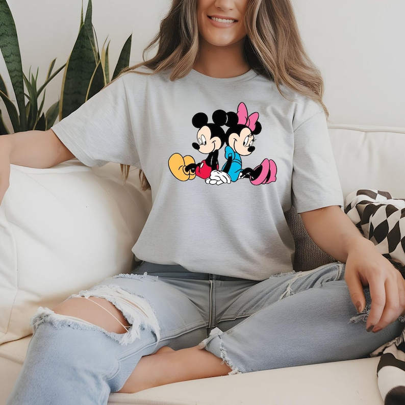 May include: Pale grey t-shirt with a colourful graphic of Mickey and Minnie Mouse. Mickey is in red shorts and yellow shoes, Minnie in a blue dress and pink shoes. The design is in a classic cartoon style, perfect for Disney fans.