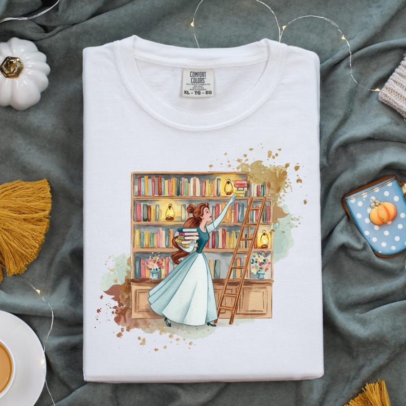 May include: White Comfort Colors t-shirt featuring a watercolor illustration of a woman in a blue dress reaching for a book on a library shelf. The image includes a wooden ladder, books, and the text "COMFORT COLORS XL - TG - EG".