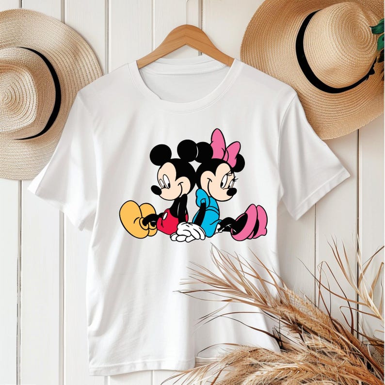 May include: White t-shirt with a cartoon graphic of Mickey and Minnie Mouse. Mickey is in red shorts and yellow shoes, Minnie in a blue dress and pink shoes. Minnie has a pink bow. The design is set against a plain white background.