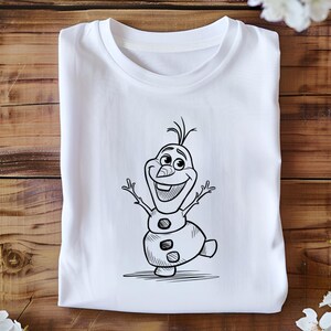 May include: White t-shirt featuring a black and white cartoon illustration of Olaf, the snowman character from the movie Frozen. Olaf is depicted with his arms raised and a cheerful expression. The t-shirt is folded neatly on a wooden surface.