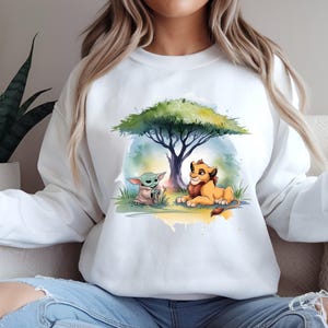 May include: White sweatshirt featuring a watercolor illustration of Baby Yoda and Simba from The Lion King under a tree. The design includes green, yellow, and blue hues. The sweatshirt is a comfortable, casual style.