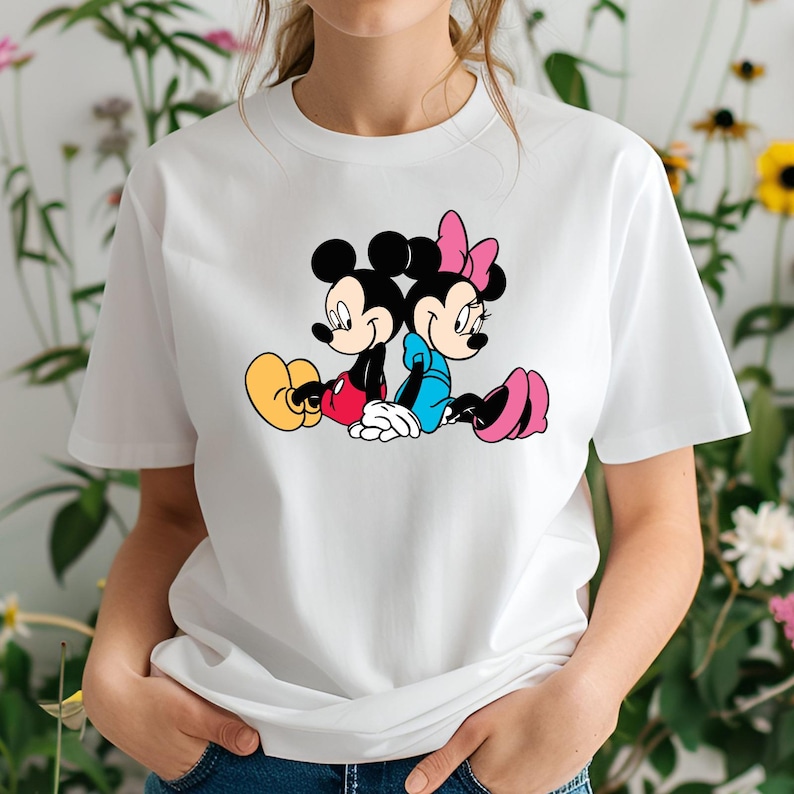 May include: White t-shirt with a colourful graphic of Mickey Mouse and Minnie Mouse. Mickey is in red shorts and yellow shoes, Minnie in a blue dress and pink shoes. The cartoon characters are depicted sitting together, leaning against each other.