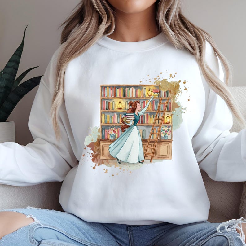 May include: White sweatshirt featuring a watercolor illustration of a woman in a blue dress reaching for a book on a bookshelf. The design includes a ladder, books, and decorative elements, creating a literary theme.
