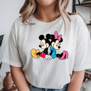 May include: White t-shirt with a cartoon illustration of Mickey Mouse and Minnie Mouse. Mickey wears red shorts and yellow shoes, Minnie a blue dress and pink shoes. The characters are sitting back-to-back. The design is set against a white background.
