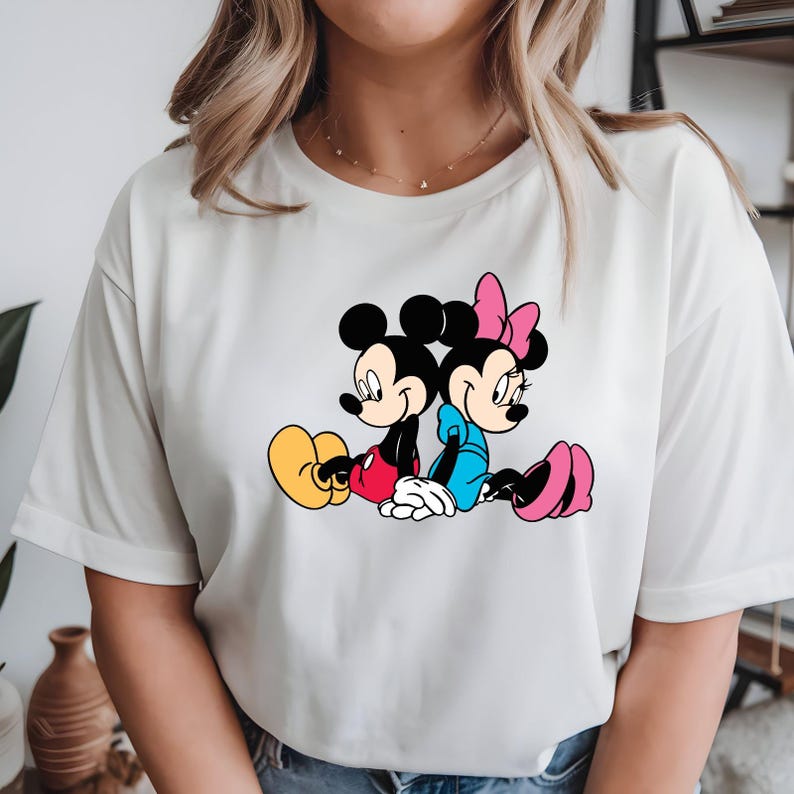 May include: White t-shirt with a cartoon illustration of Mickey Mouse and Minnie Mouse. Mickey wears red shorts and yellow shoes, Minnie a blue dress and pink shoes. The characters are sitting back-to-back. The design is set against a white background.