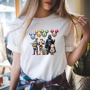 Puede incluir: Camiseta blanca con un gr&aacute;fico colorido de personajes de Star Wars y globos de Mickey Mouse. El dise&ntilde;o incluye a Darth Vader, The Mandalorian, Grogu y otros personajes. Los globos son de varios colores.