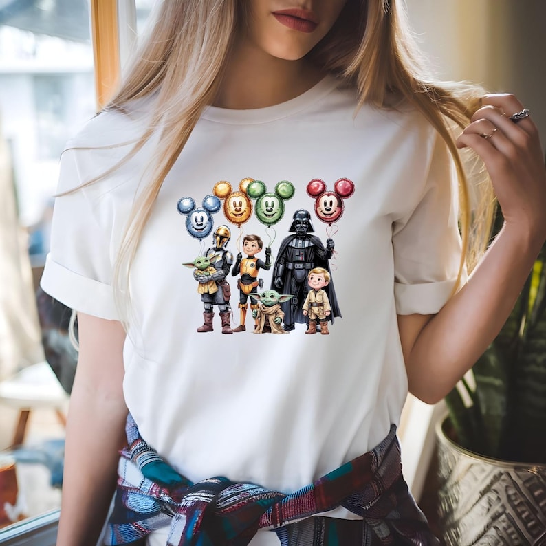 Puede incluir: Camiseta blanca con un gr&aacute;fico colorido de personajes de Star Wars y globos de Mickey Mouse. El dise&ntilde;o incluye a Darth Vader, The Mandalorian, Grogu y otros personajes. Los globos son de varios colores.