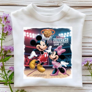 May include: White crew neck sweatshirt featuring a colorful graphic of Mickey and Minnie Mouse playing basketball. The graphic includes the text "Disney Dunkers Game On!" Mickey Mouse is wearing a red and yellow basketball uniform. Minnie Mouse is wearing a pink outfit.