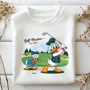Könnte beinhalten: Weißes Sweatshirt mit einer Cartoon-Illustration von Donald Duck beim Golfspielen. Das Design enthält den Text "Golf Masters - TEE TIME!" und zeigt Donald Duck in Golfkleidung auf einem grünen Golfplatz. Ein zweiter Donald Duck ist ebenfalls vorhanden.