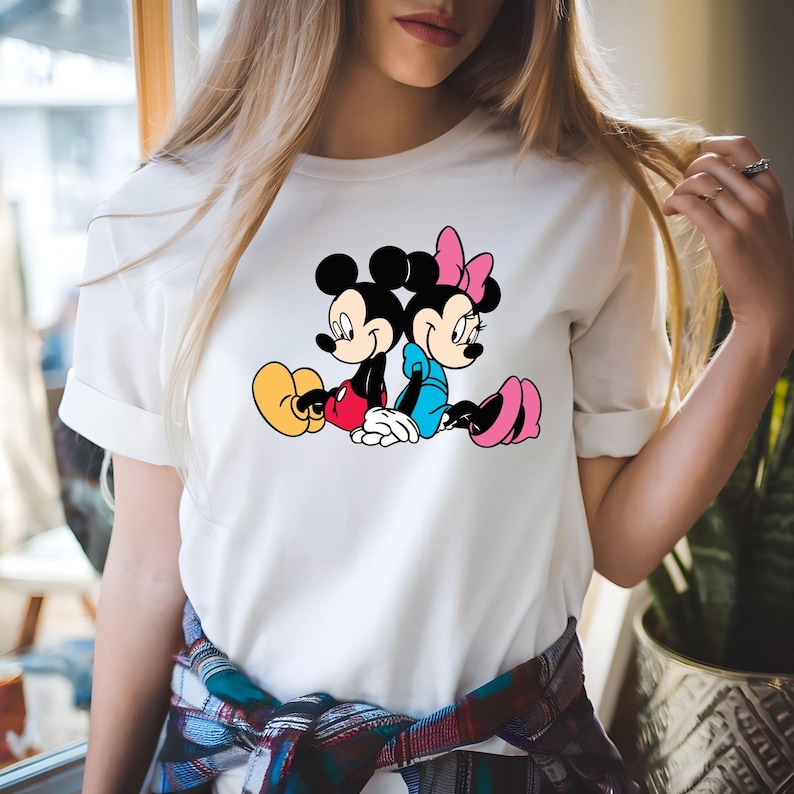 May include: White t-shirt with a colourful graphic of Mickey and Minnie Mouse sitting back-to-back. Mickey wears red shorts and yellow shoes, while Minnie is in a blue dress and pink shoes. The cartoon characters are set against a white background.