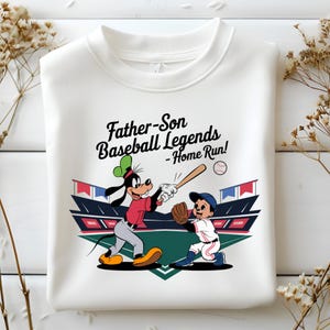 May include: White sweatshirt with a baseball graphic and the text "Father-Son Baseball Legends - Home Run!". The design shows a cartoon of Goofy batting and a boy in a baseball uniform, set against a stadium backdrop.