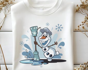 Puede incluir: Sudadera blanca con un dibujo animado de Olaf sosteniendo una escoba. El diseño incluye salpicaduras de agua azules y copos de nieve. Olaf lleva un sombrero y tiene una nariz de zanahoria. La sudadera es de cuello redondo.