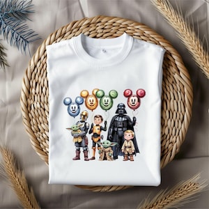 Puede incluir: Camiseta blanca de cuello redondo con un gr&aacute;fico colorido de personajes de Star Wars y globos con forma de Mickey Mouse. El dise&ntilde;o incluye a Darth Vader, Grogu y otros personajes. La camiseta se muestra sobre una superficie tejida.