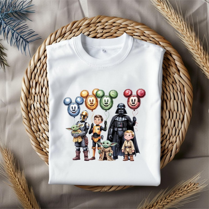 Puede incluir: Camiseta blanca de cuello redondo con un gr&aacute;fico colorido de personajes de Star Wars y globos con forma de Mickey Mouse. El dise&ntilde;o incluye a Darth Vader, Grogu y otros personajes. La camiseta se muestra sobre una superficie tejida.