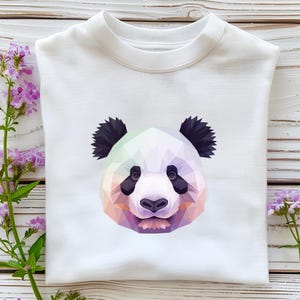 May include: White sweatshirt featuring a geometric panda face design. The panda's face is composed of various pastel colors, with black ears and eyes. The sweatshirt is folded neatly on a light wooden surface, with purple flowers in the background.