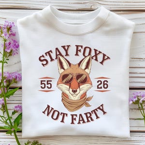 May include: White sweatshirt with a graphic of a fox wearing sunglasses and a bandana. The text reads "STAY FOXY NOT FARTY" with the numbers 55 and 26 on either side of the fox. The sweatshirt is folded.