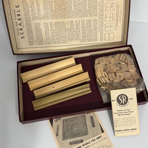 May include: An open Scrabble game box with wooden letter holders and tiles. The box contains game instructions and a pamphlet with the text "What Word Would You Add?" The game is from Selchow & Righter Company.