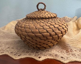 May include: A woven basket with a lid, crafted from natural fibers in shades of brown and tan. The basket has a rounded shape with a lid and a small handle on top. The basket is sitting on a cream-colored, open-weave fabric.