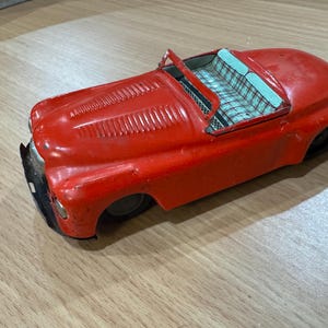May include: A vintage red toy car with a convertible top. The car has a classic design with a detailed front grill and a plaid interior. The toy car is made of metal and shows signs of wear, suggesting it is an antique.