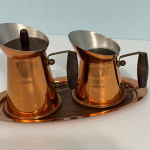 May include: A copper-coloured creamer and sugar set with wooden handles and an oval tray. The creamer has a silver-coloured interior and a wooden lid. The set is designed for serving coffee or tea.