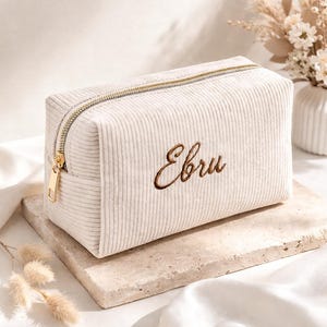 Könnte beinhalten: Ein rechteckiges, cremeweißes Etui aus Kordstoff mit dem in brauner Schreibschrift gestickten Namen Ebru auf der Vorderseite. Das Accessoire verfügt über einen goldfarbenen Metallreißverschluss und eine gerippte Stoffoberfläche.