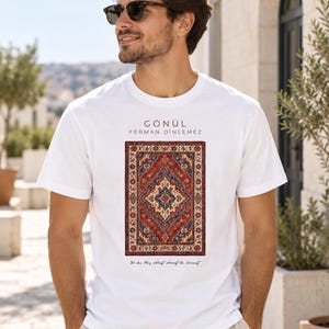 Puede incluir: Una camiseta blanca de manga corta con un gráfico central de una alfombra rectangular con intrincados patrones geométricos en rojo, azul y crema. Sobre la alfombra, el texto GÖNÜL FERMAN DİNLEMEZ y debajo, Wo das Herz schlägt, schweigt die Vernunft.