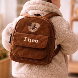 Könnte beinhalten: Ein brauner Kordrucksack mit einem kleinen gestickten Welpengesicht im oberen Bereich und dem Namen Theo in weißem Garn auf der Vordertaschenklappe. Die Tasche verfügt über verstellbare Schultergurte und einen oberen Tragegriff.