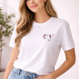 Könnte beinhalten: Weißes T-Shirt mit floraler Stickerei "ey" auf der Brust. Das Shirt wird mit hellblauen Jeans kombiniert. Das Model trägt eine goldene Halskette und hat lange blonde Haare.