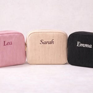 Könnte beinhalten: Drei Kosmetiktaschen aus Cord in Rosa, Beige und Schwarz. Jede Tasche hat einen goldenen Reißverschluss und einen aufgestickten Namen auf der Vorderseite: Lea, Sarah und Emma. Die Taschen sind rechteckig mit abgerundeten Ecken.