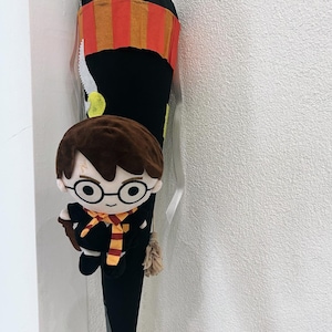 Puede incluir: Una bolsa escolar negra en forma de cono con un muñeco de peluche de Harry Potter adjunto. La bolsa tiene una sección a rayas rojas y naranjas cerca de la parte superior. El muñeco tiene pelo castaño, gafas y una bufanda de Gryffindor.