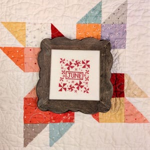 May include: A square cross-stitch embroidery piece featuring the word JUNE in red thread, surrounded by small red pinwheel motifs. The embroidery is displayed in a dark brown wooden frame with a scalloped edge, centered on a white quilted fabric.