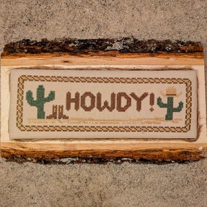 May include: A cross-stitched sign on a wooden plaque with the word "HOWDY!" in brown letters. The design includes a cactus, cowboy boots, and a cactus wearing a hat. The sign is framed with a decorative border.