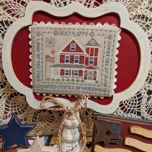 May include: A framed cross-stitch featuring a red and white house with a flag, and the text "July 4, 1776" and "Home of the Brave." The frame is white with a scalloped edge, set against a red background. Decorative stars and a wooden flag are in the foreground.