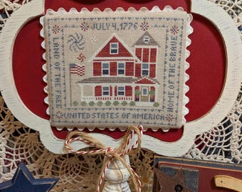 May include: A framed cross-stitch featuring a red and white house with the text "July 4, 1776", "Land of the Free", "Home of the Brave", and "United States of America". The frame is white with a decorative edge, set against a red background.