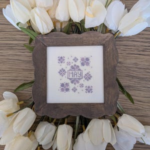 May include: A framed cross-stitch piece with the word "MAY" in the center, surrounded by floral patterns in shades of purple. The frame is a dark brown, and the piece is set against a backdrop of white tulips and a wooden surface.