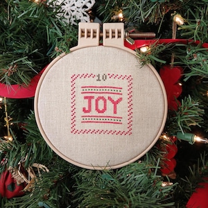 May include: A round, beige embroidery hoop with the word "JOY" stitched in red thread. The embroidery is framed by a red cross-stitch border and the number "10" is stitched above the word. The hoop is hanging on a Christmas tree.