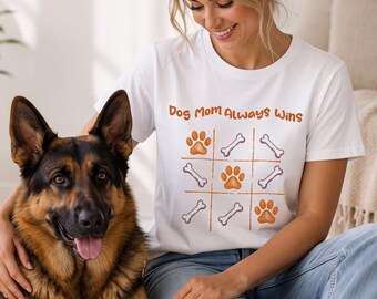 May include: A white short-sleeved t-shirt featuring a graphic of a tic-tac-toe board with orange paw prints and white bones. The text "Dog Mom Always Wins" is printed in orange above the game board design on the front of the garment.