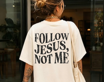 Könnte beinhalten: Ein elfenbeinfarbenes Kurzarm-T-Shirt mit dem schwarzen, abgenutzten Schriftzug "FOLLOW JESUS, NOT ME" auf der Rückseite. Das Shirt hat einen Rundhalsausschnitt und eine lockere Passform. Eine passende elfenbeinfarbene Stofftasche wird über der Schulter getragen.