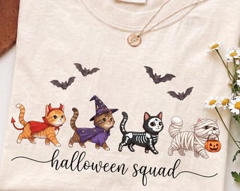 Könnte beinhalten: Ein elfenbeinfarbenes T-Shirt mit einer Grafik von vier Katzen in Halloween-Kostümen: ein Teufel, eine Hexe, ein Skelett und eine Mumie. Über den Katzen sind drei kleine Fledermäuse und darunter der Text "halloween squad" in schwarzer Schrift.