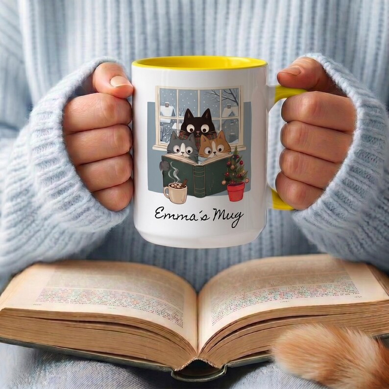 May include: A white ceramic mug with a yellow interior and handle, featuring a graphic of three cats reading a book by a window, a small potted plant, and a cup of cocoa. The text "Emma's Mug" is printed in black script below the illustration.