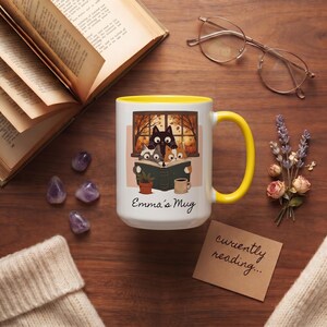 May include: A white ceramic mug with a bright yellow interior and handle. The mug features a graphic of three cats reading a book by a window with autumn leaves. Below the illustration, the text reads Emma's Mug in a black cursive script.
